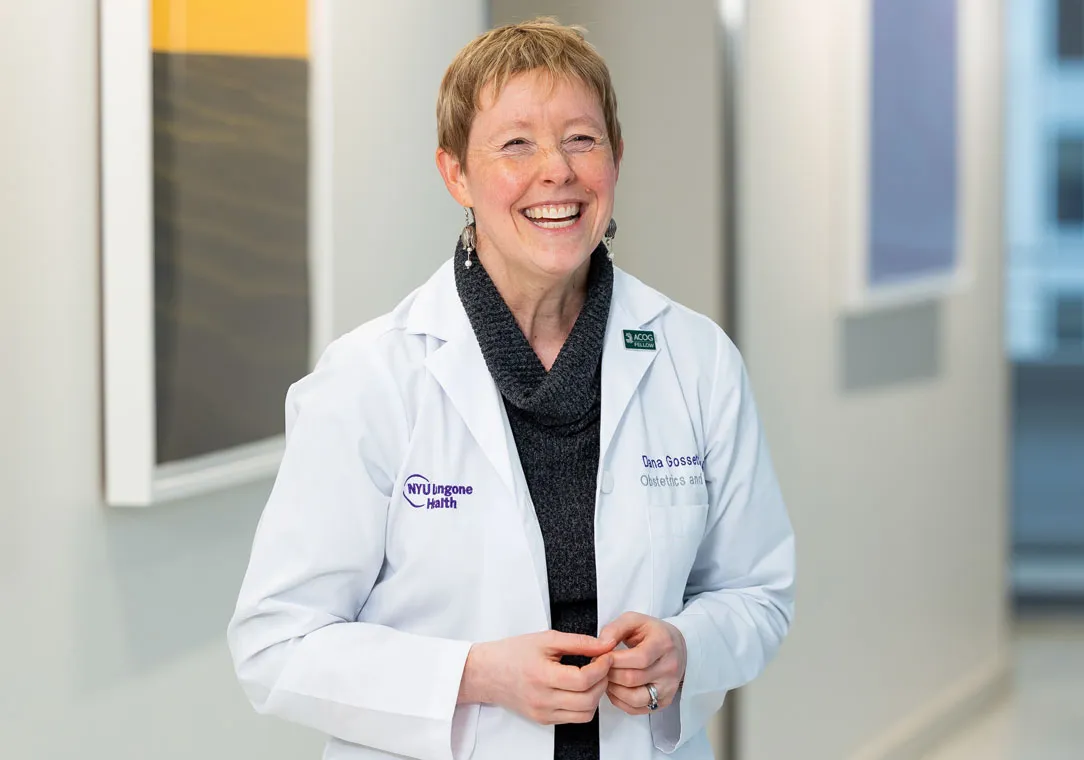 Dr. Dana Gossett stands smiling in a well-lit hallway with framed artwork on the wall in the background.