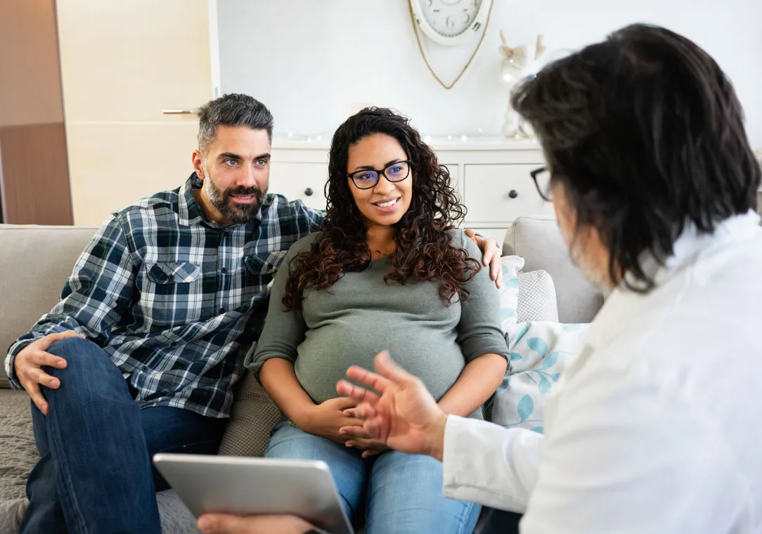 A man and pregnant woman sit on a couch while talking to a healthcare provider. 