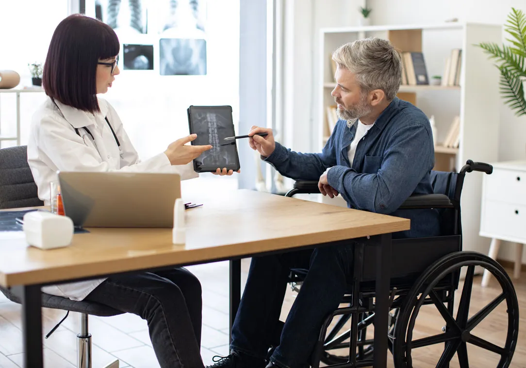 A doctor discusses X-ray results with a patient sitting in a wheelchair at a desk in a medical office.