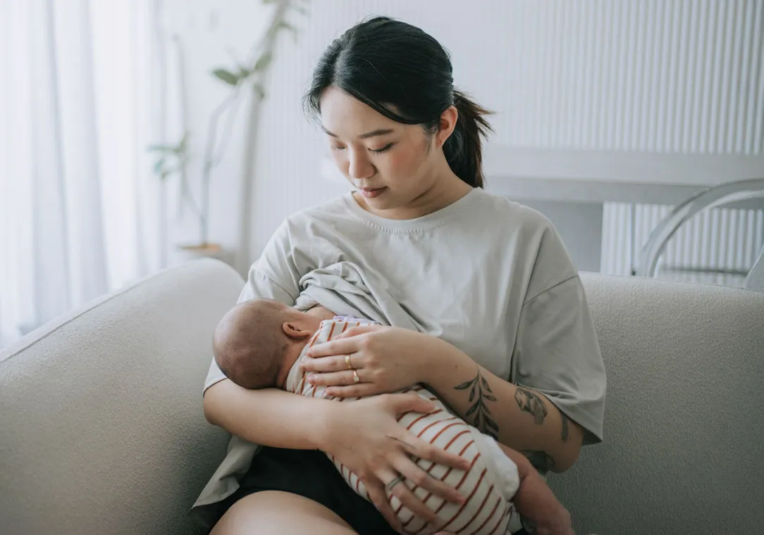 A mother sits on a couch while breastfeeding her baby. 