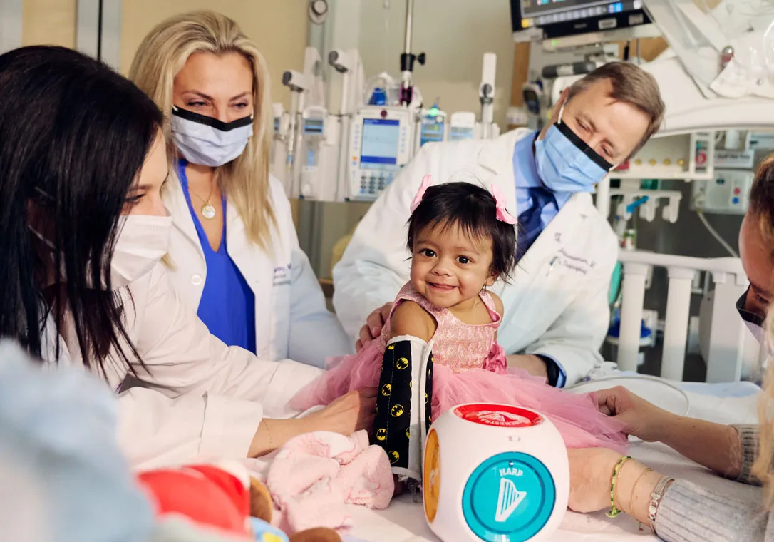 Two medical professionals wearing lab coats and face masks examining an infant child in a hospital room with toys scattered around the foreground.