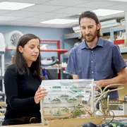 Two people examine a technological prototype in a lab setting, surrounded by equipment and tools.