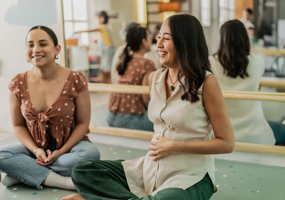 Two women sit cross-legged in a room with a large mirror on the back wall; one is pregnant, resting her hand on her belly.