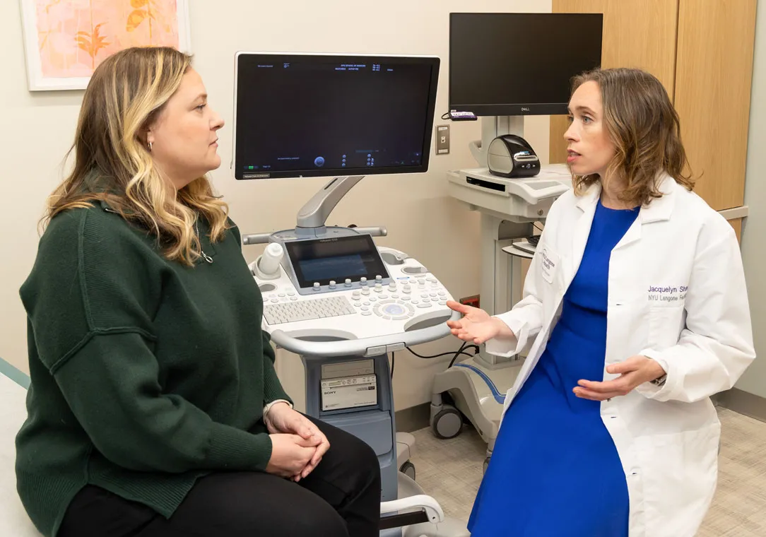 A woman sits in a medical office talking with Dr. Jacquelyn Shaw, with ultrasound equipment nearby.