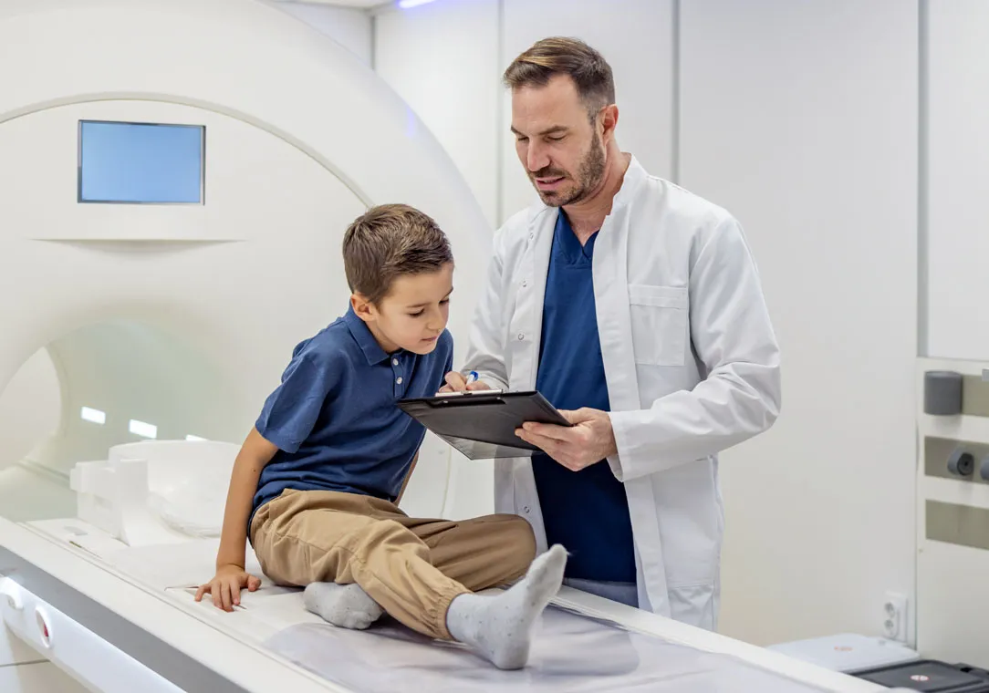 A child sits on an MRI machine while a healthcare provider reviews information on a tablet in a diagnostic imaging room.
