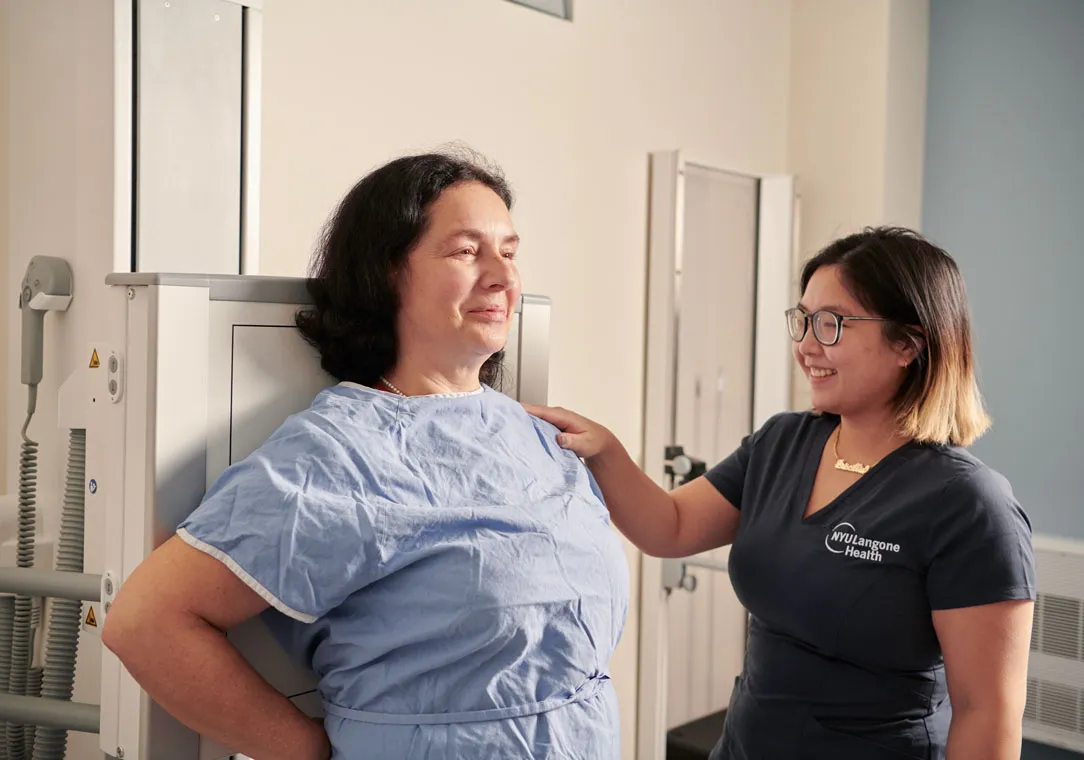 A healthcare worker helps a patient in a medical gown adjust their position for an imaging scan in a clinical examination room.