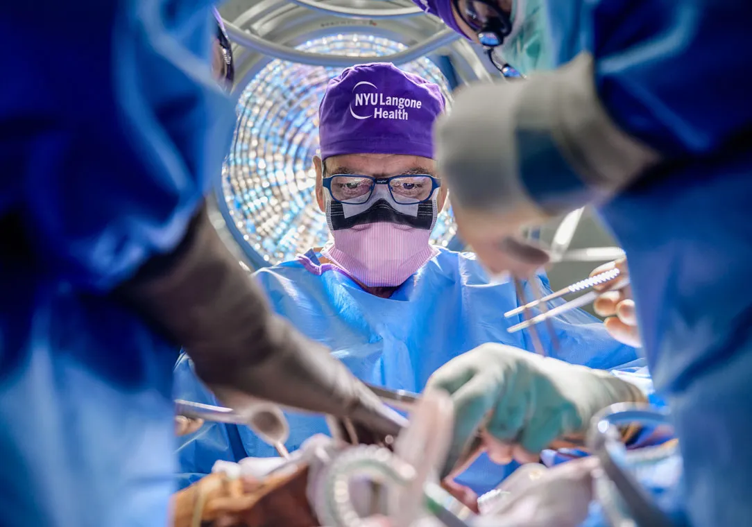 A surgeon wearing full personal protective equipment, surgical cap, face mask, and safety glasses stands over an operating table. Other unidhttps://clinicaltrials.med.nyu.edu/clinicaltrials/search/?q=liver%20transplantentifiable medical staff stand obscured in the foreground.