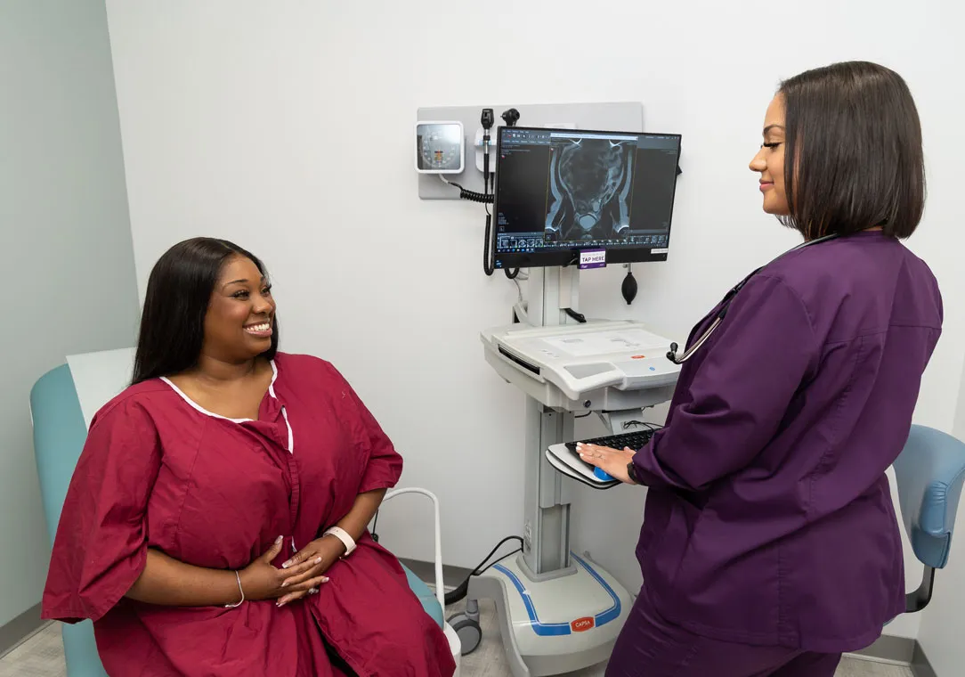 A sonographer stands in front of ultrasound equipment while speaking to a patient in an exam room.