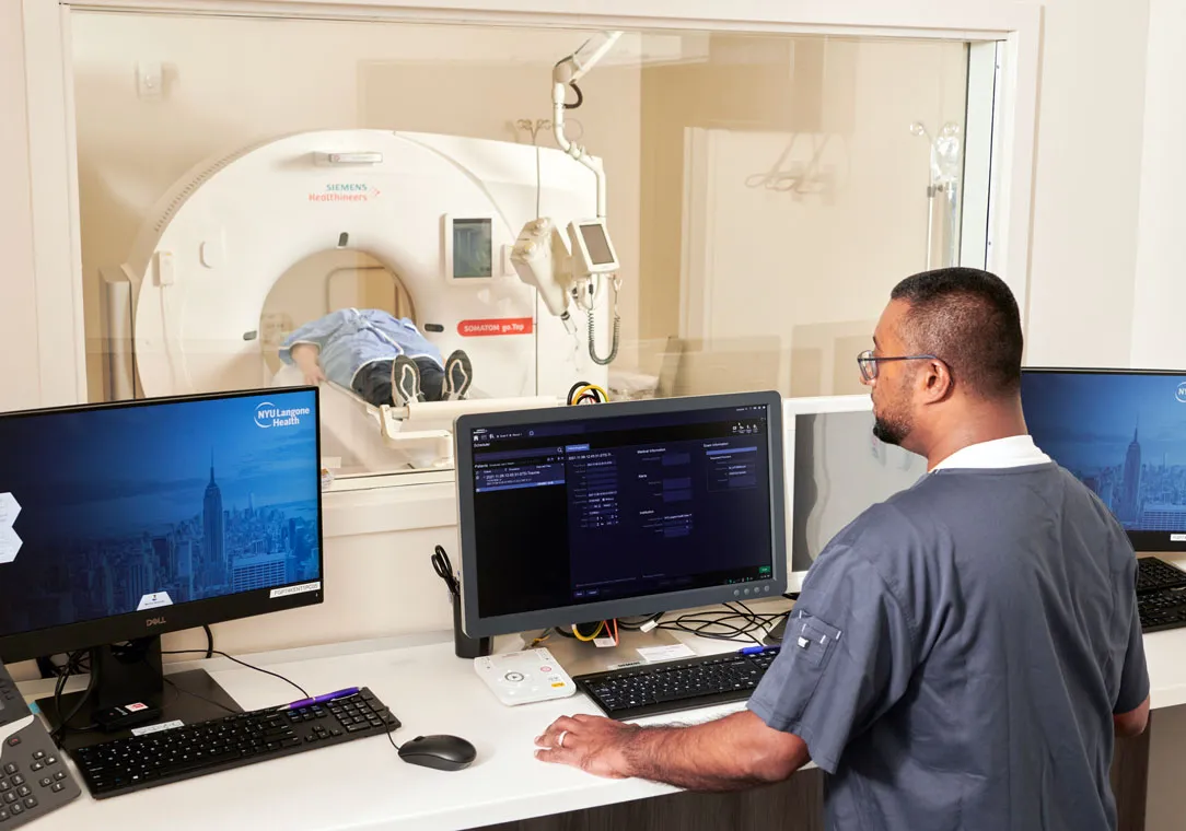 A technician monitors a patient undergoing a CT scan in an imaging room, with computers displaying diagnostic software.