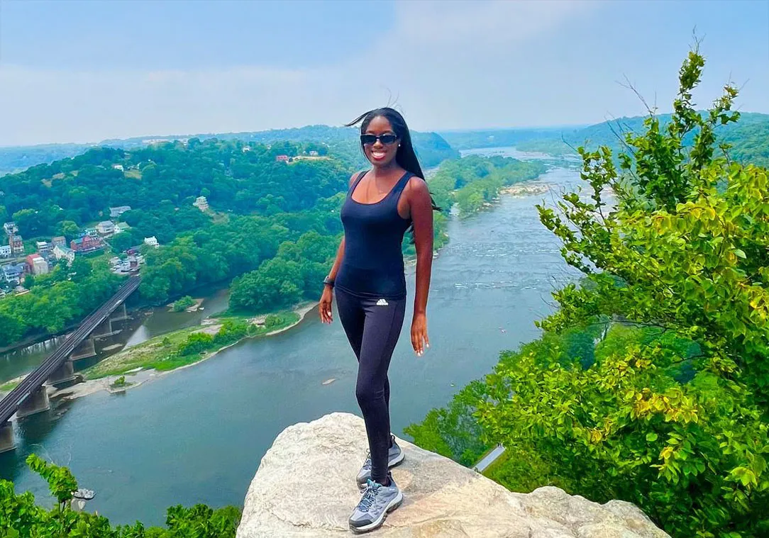 A woman in athletic wear stands on a rock overlooking a river valley with lush greenery.