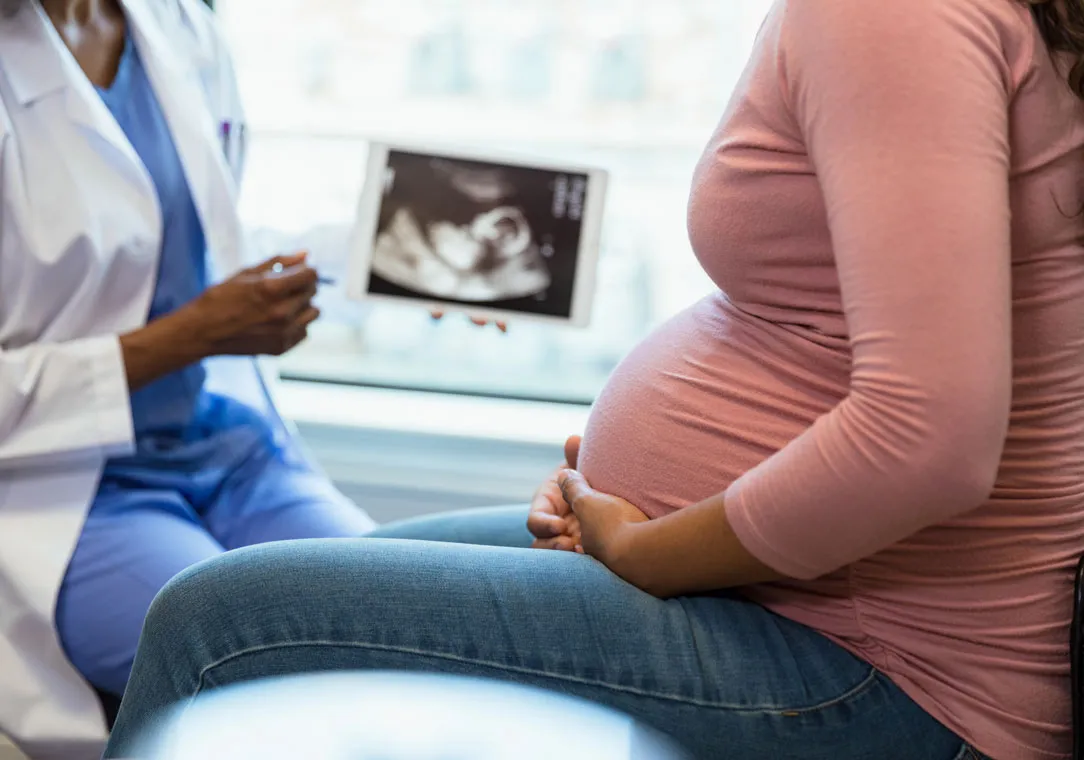 A pregnant person sits, facing a doctor holding an ultrasound image, both partially visible in a clinic setting.