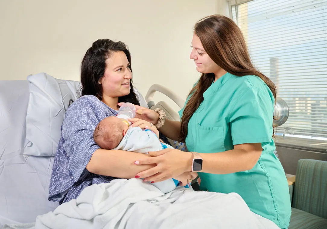 Nurse Alexandra Minucci helps a mother sitting in a hospital bed bottle-feed her baby.