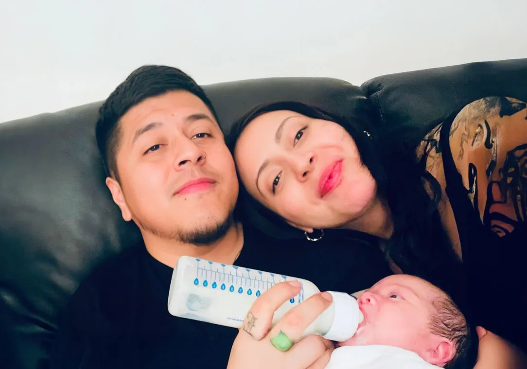 A man and a woman sit on a couch while bottle-feeding their baby.