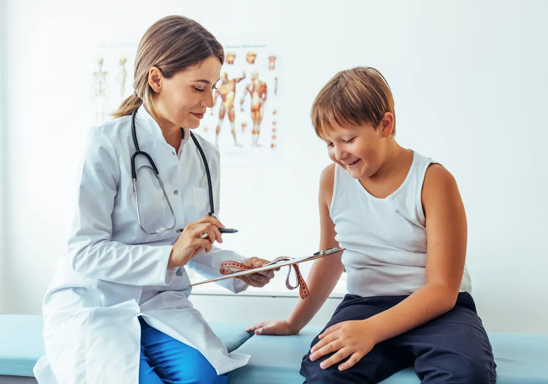 A healthcare provider holds a measuring tape and a clipboard as she talks with a young patient. 