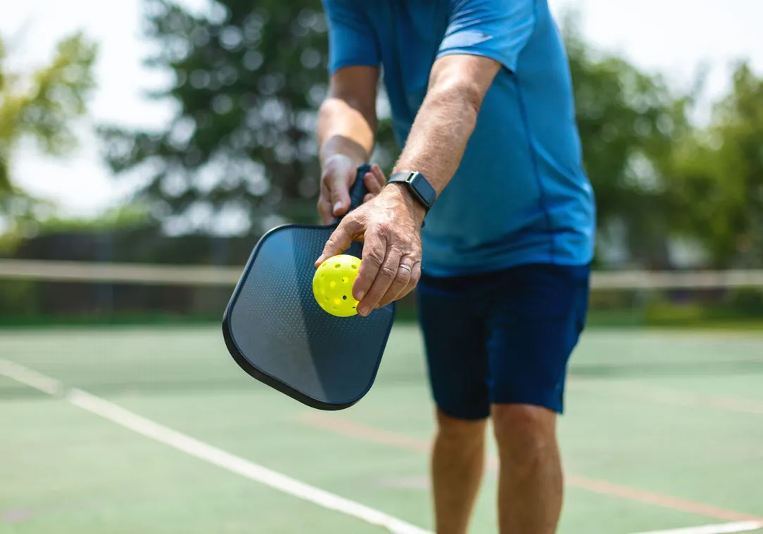 A person playing pickleball outdoors holds a paddle and a yellow ball on a court surrounded by trees.