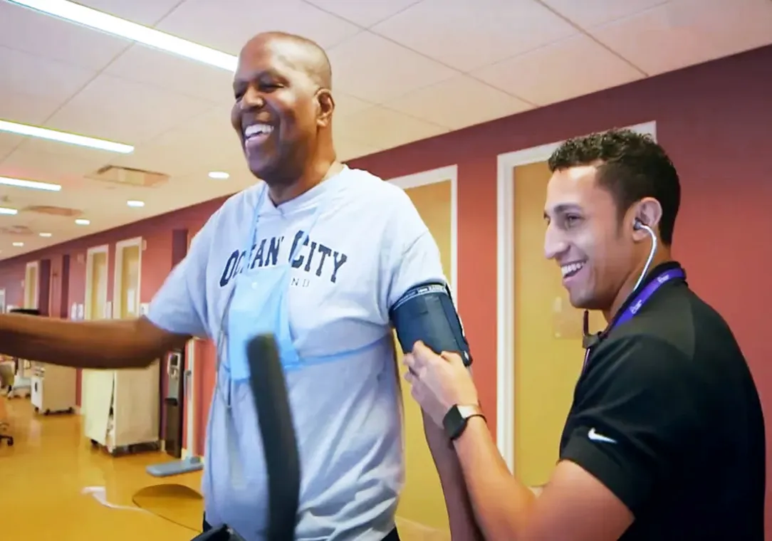A man wearing a gray shirt smiles while standing on a piece of exercise equipment as a medical professional takes his blood pressure.