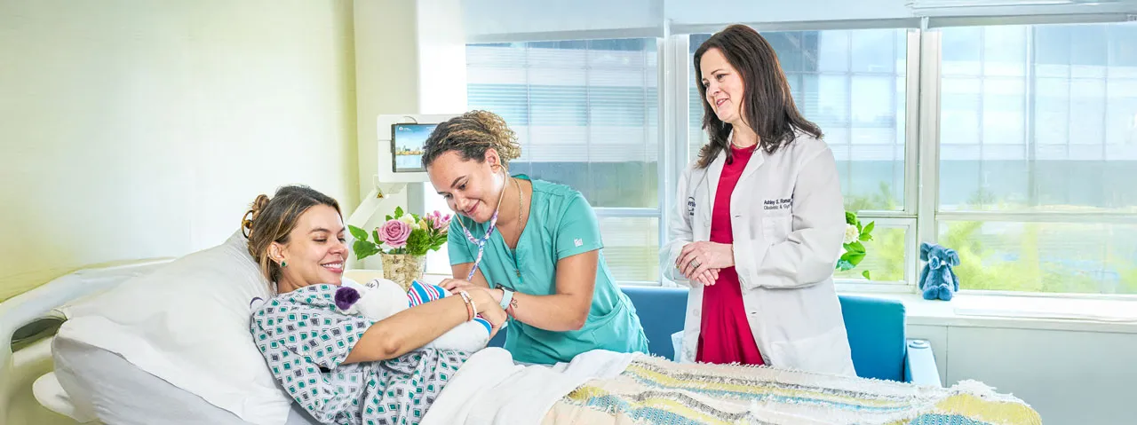 Dr. Ashley Roman and nurse Jordana Reyes stand at the bedside of a woman holding a newborn baby.