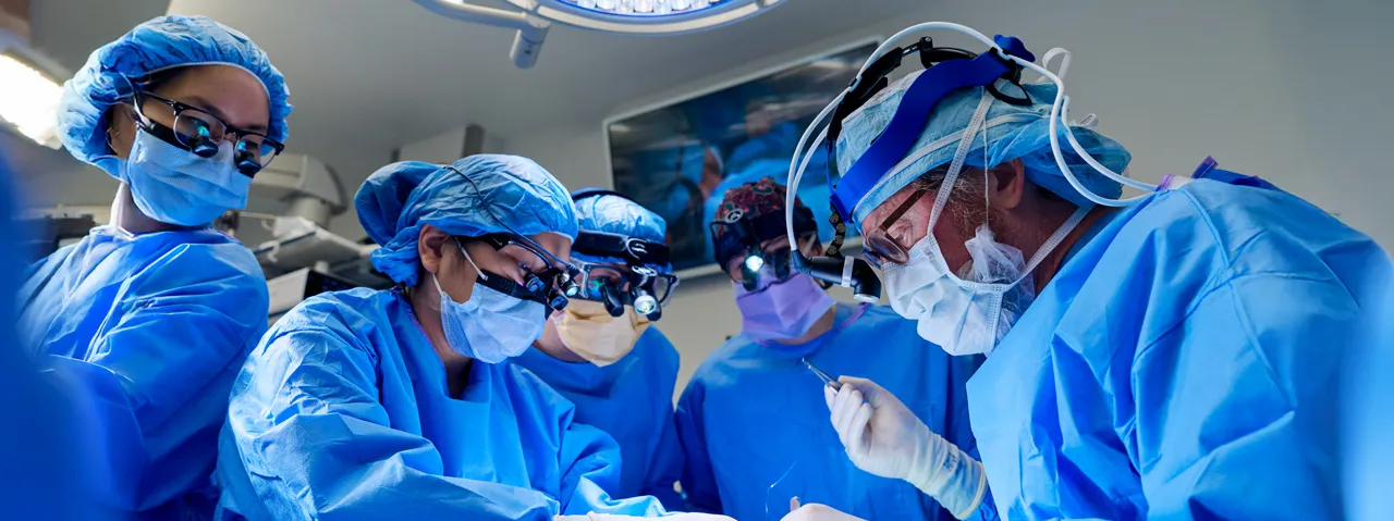 A team of five medical professionals wearing personal protective equipment stand around an operating table during a medical procedure.