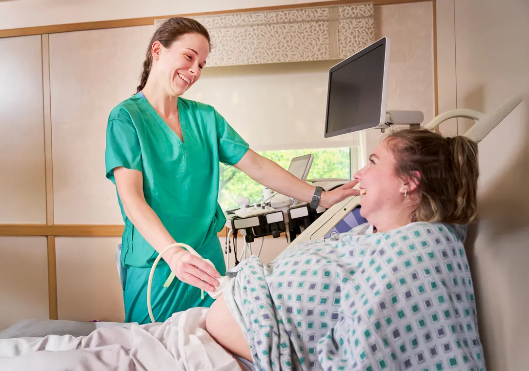  Dr. Samantha Hill, wearing scrubs, adjusts monitoring equipment while talking with a patient lying in a hospital bed.  