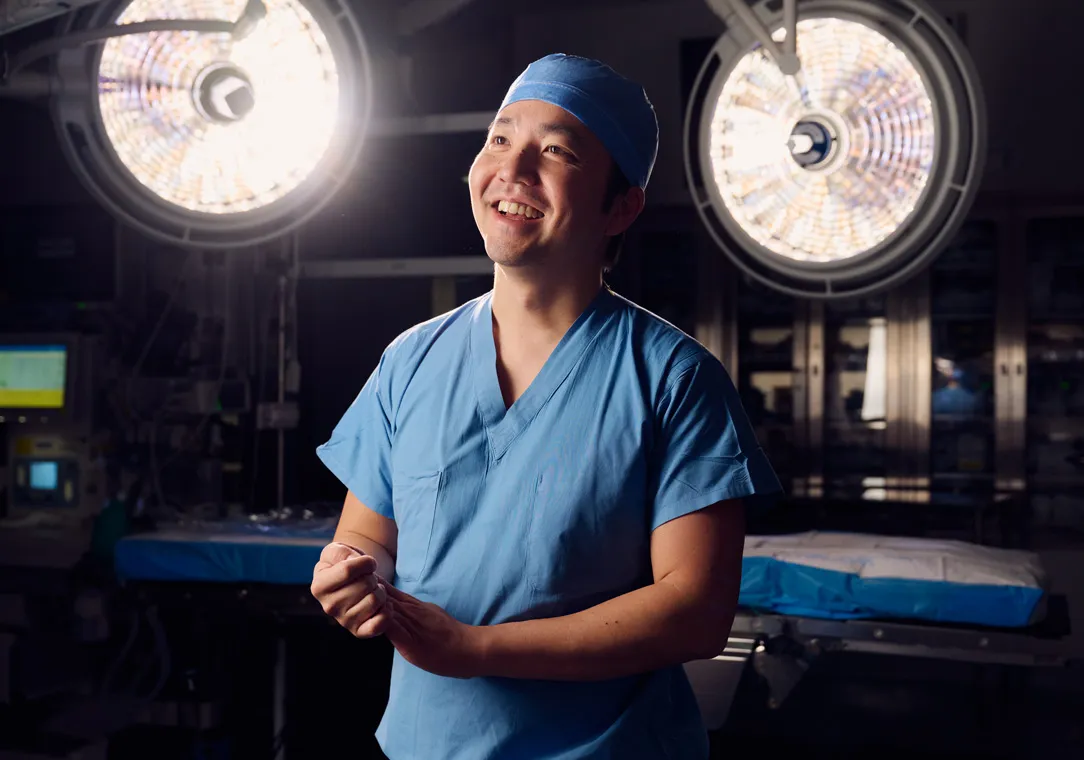 Dr. Jun Tashiro wears scrubs while standing in an operating room with bright surgical lights in the background.