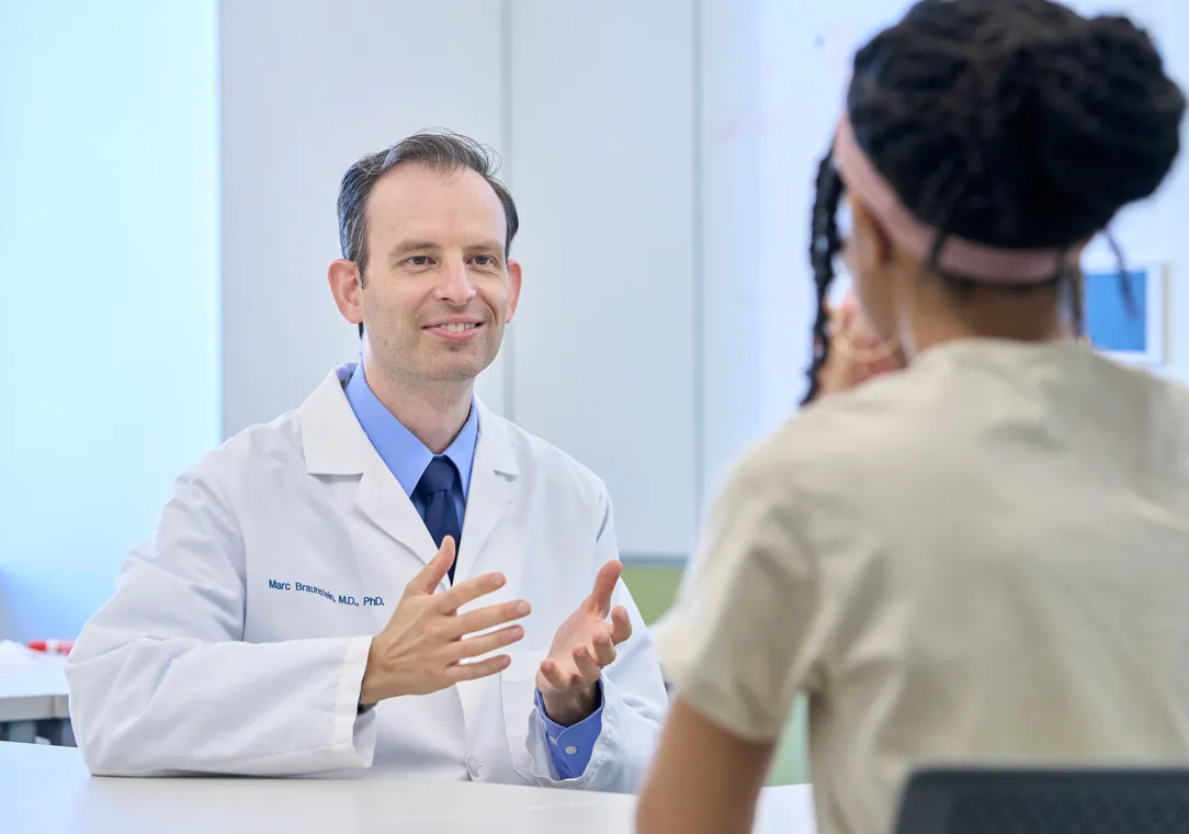 A doctor in a white lab coat sits at a desk, gesturing with his hands while speaking to a patient seated across from him in a clinical office setting.